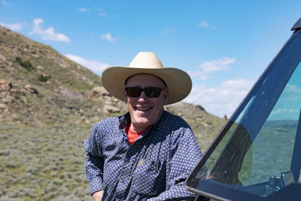 A smiling man in a cowboy hat and sunglasses stands outdoors by a vehicle on a cattle ranch, with hills and blue sky in the background.