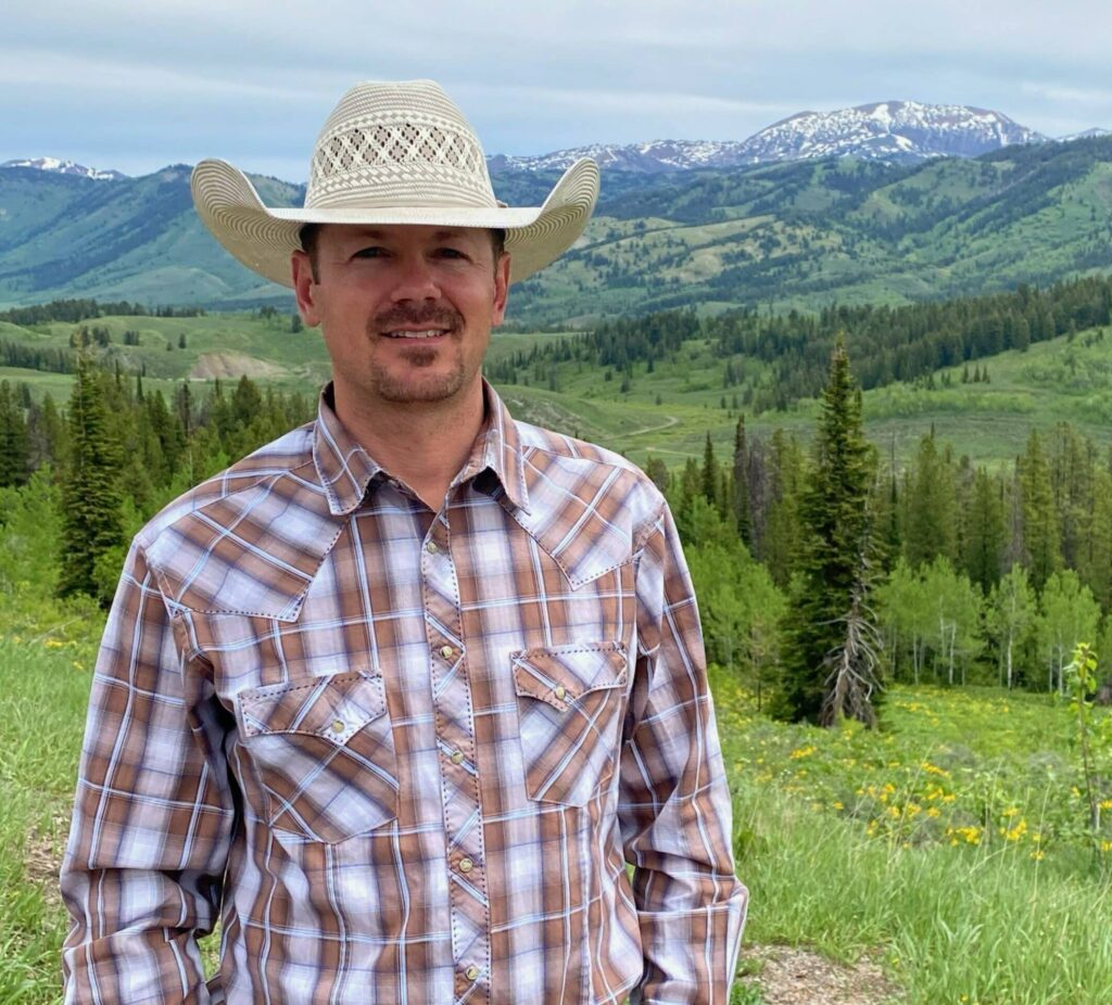 A man in a straw cowboy hat and plaid shirt stands outdoors on a green cattle ranch, set against a mountainous landscape with scattered trees and snow-capped peaks.