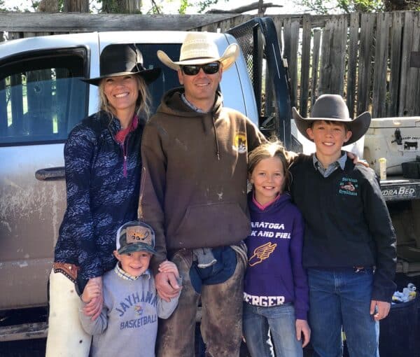A smiling family of five in cowboy hats and casual clothes stands together outdoors on recreational land in front of a truck and wooden fence, with sunshine casting shadows around them.
