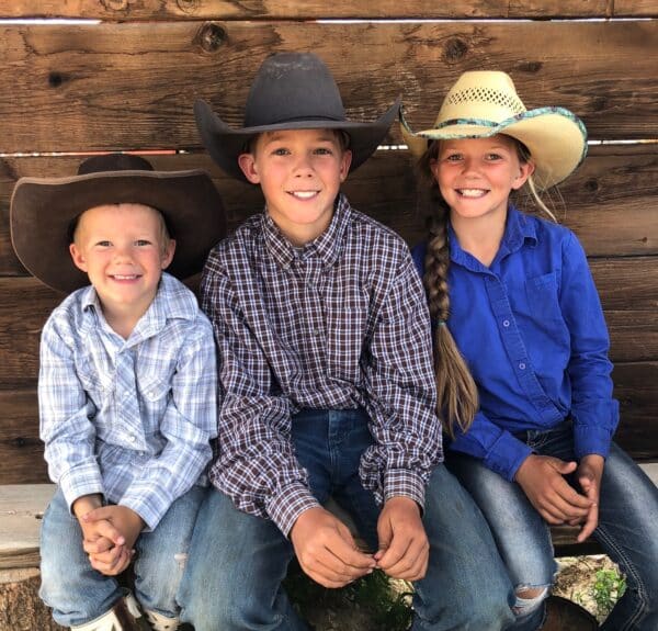 Three smiling children wearing cowboy hats, plaid shirts, and jeans sit on a wooden bench in front of a rustic wall—perfectly capturing the spirit of life on a cattle ranch. The two boys are on the left and center; the girl with a long braid sits on the right.