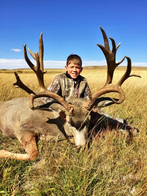 A smiling boy in a camouflage jacket poses behind a large deer with impressive antlers, sitting in tall grass under a clear blue sky—showcasing the excitement of recreational land on a beautiful open field.
