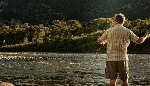 A man stands in a river while fly fishing on scenic recreational land, casting a line with trees and forested hills in the background under warm sunlight.