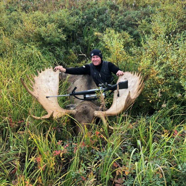 A person dressed in black sits behind a large moose with wide antlers lying in tall grass, holding its antlers and smiling at the camera. A rifle rests across the moose’s antlers, surrounded by dense green vegetation on prime hunting property.