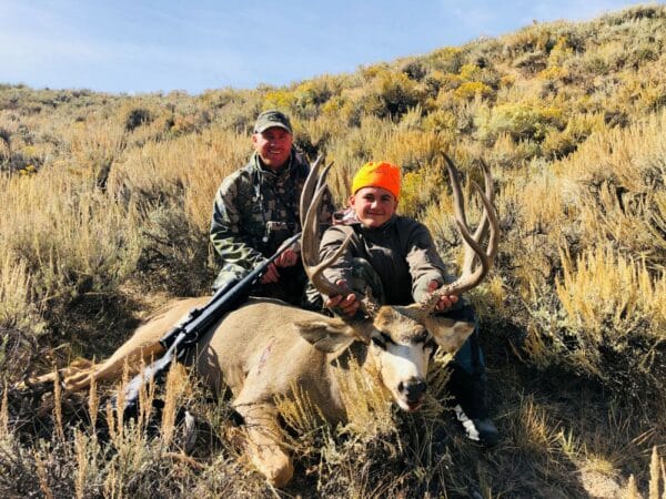 Two people outdoors in tall grass on recreational land pose with a large deer they have hunted; one wears camouflage and holds a rifle, while the other, in an orange hat, holds the deer’s antlers.
