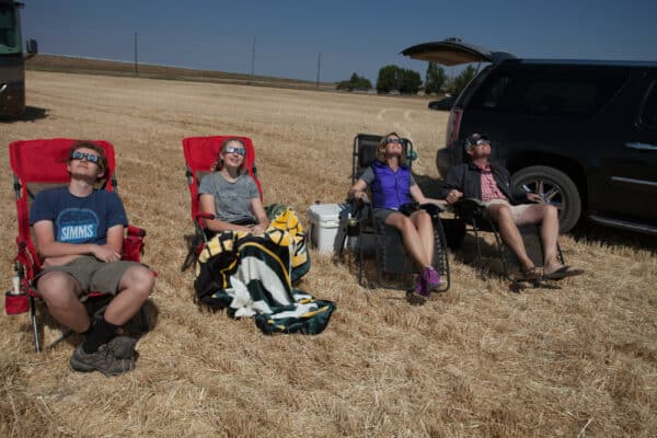 Four people sit on chairs in a dry, grassy field next to a black SUV on open land for sale, wearing solar eclipse glasses and looking up at the sky. Two are wrapped in a blanket. It is sunny and clear, with open fields in the background.