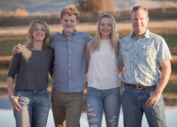 A family of four stands outdoors in casual clothes, smiling and posing together on a grassy field with a pond in the background—perfect for those seeking hunting property or land for sale.
