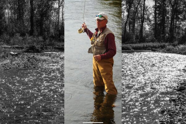 A person wearing waders, a vest, and a cap stands fly fishing in a river on recreational land for sale. The central area is in color, while the surroundings are black and white.