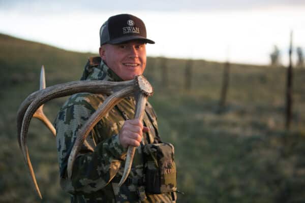 A smiling person in camouflage gear and a cap holds a large deer antler over their shoulder while standing outdoors on hunting property near a fence at sunset.