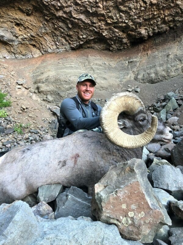 A smiling man in outdoor gear and a camo cap poses with a large bighorn sheep, surrounded by rocks and a rugged cliff—an ideal scene on prime hunting property.