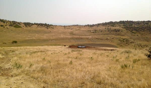 A dry, grassy landscape with gently rolling hills under a hazy sky—ideal as a hunting property or cattle ranch. In the middle distance sits a small, circular water trough surrounded by bare earth. Sparse shrubs and rocks dot the land for sale.