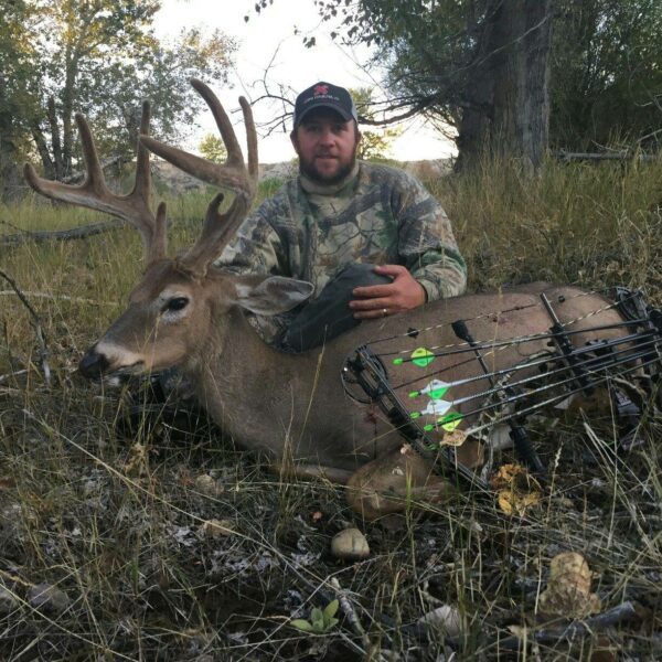 A man in camouflage poses outdoors with a large deer he has hunted, resting his hand on the deer's body. A compound bow with green-fletched arrows lies across the deer. Trees and grass hint at premium hunting property in the background.