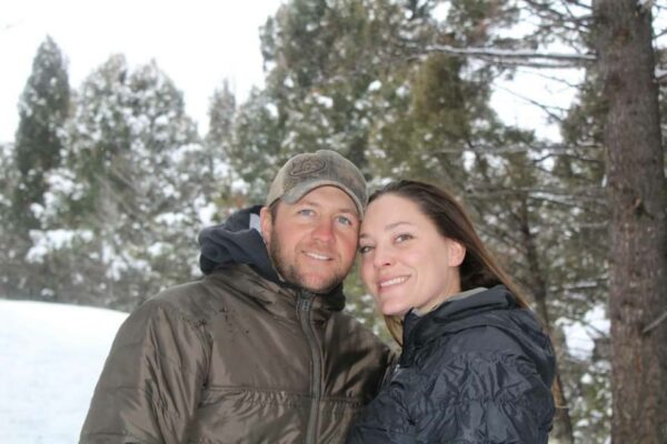 A man and woman, both in winter jackets, stand close together and smile in a snowy forest of tall evergreens, enjoying the beauty of recreational land.