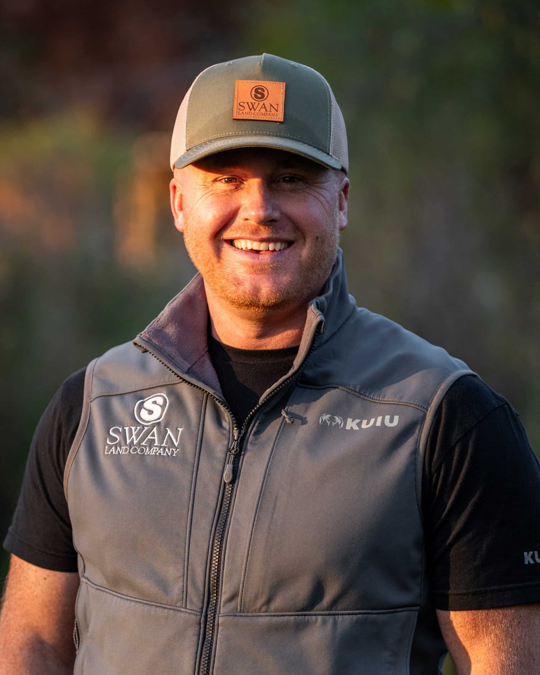 A smiling man wearing a gray cap and a gray sleeveless vest with Swan Land Company and KUIU logos stands outdoors in natural sunlight, representing recreational land and hunting property.