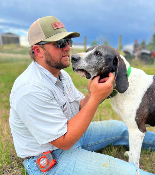A man in sunglasses, a cap, and a light shirt sits on grass, gently holding the face of a large black and white dog with a green collar. The outdoor scene hints at life on a cattle ranch under a cloudy sky.