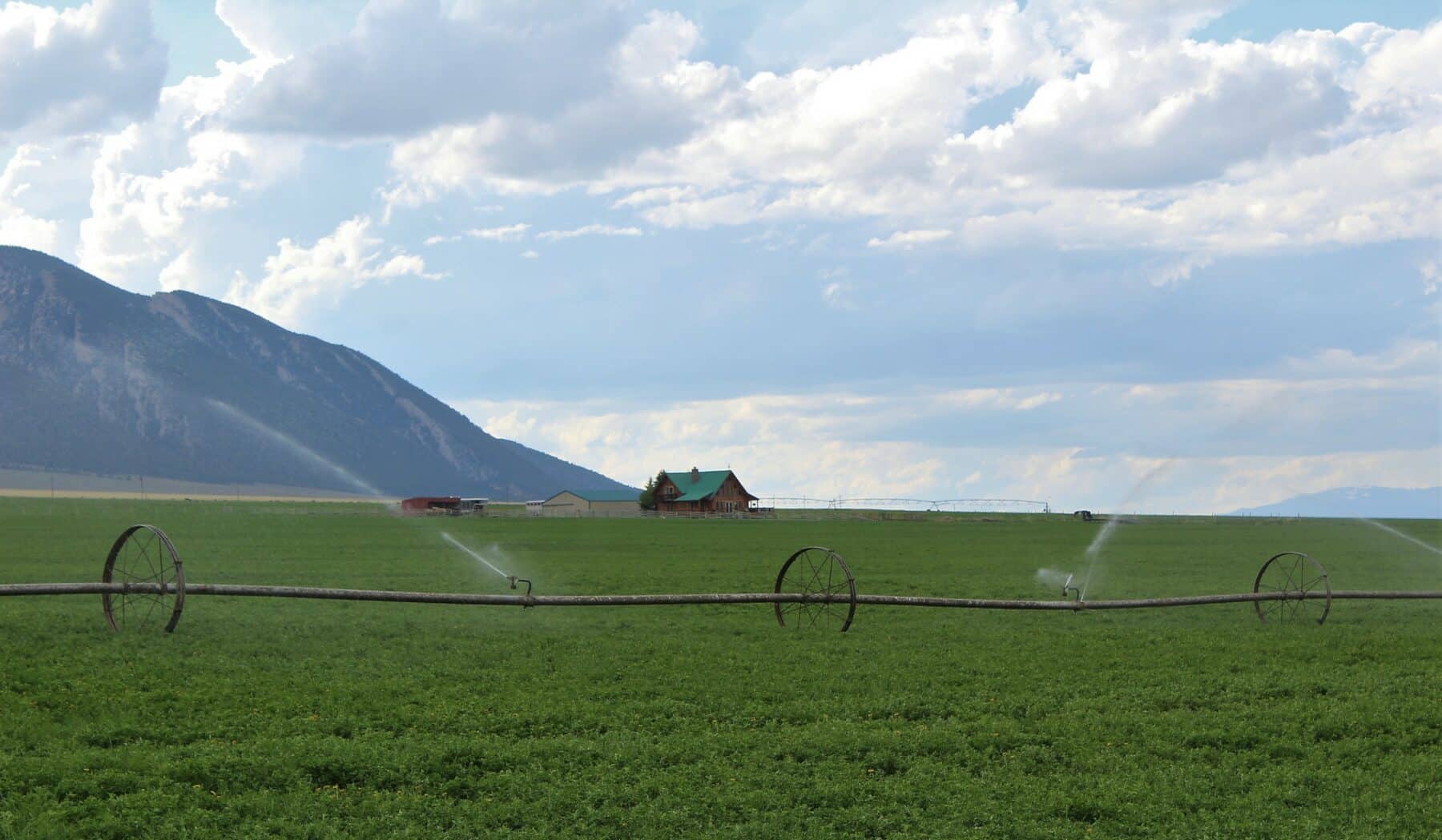 A lush green field is being irrigated by sprinklers, with a farmhouse and red barn in the distance—ideal as a cattle ranch or land for sale. Majestic mountains rise in the background under a partly cloudy sky.