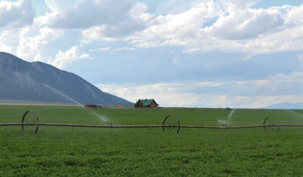 A lush green field is being irrigated by sprinklers, with a farmhouse and red barn in the distance—ideal as a cattle ranch or land for sale. Majestic mountains rise in the background under a partly cloudy sky.