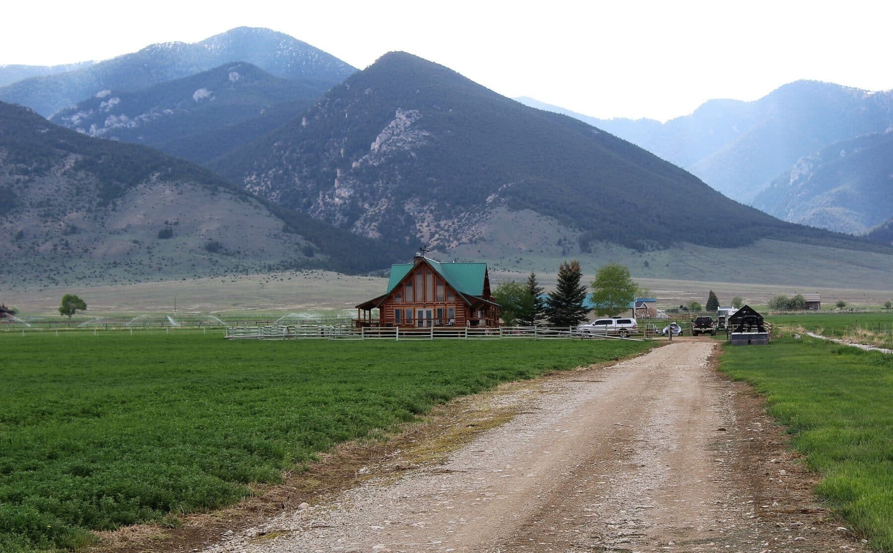 A gravel road leads to a wooden house with a green roof on recreational land, surrounded by fields and trees, set against tall, forested mountains under a partly cloudy sky.