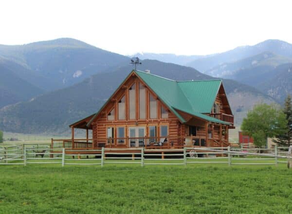 A large wooden log cabin with a green metal roof sits on recreational land in front of tree-covered mountains, surrounded by a wooden fence and lush grass under a cloudy sky.
