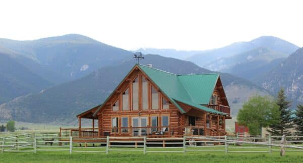 A charming wooden log cabin with a green roof sits behind a fence on recreational land, surrounded by green grass, trees, and tall mountains beneath a partly cloudy sky—a perfect ranch for sale.