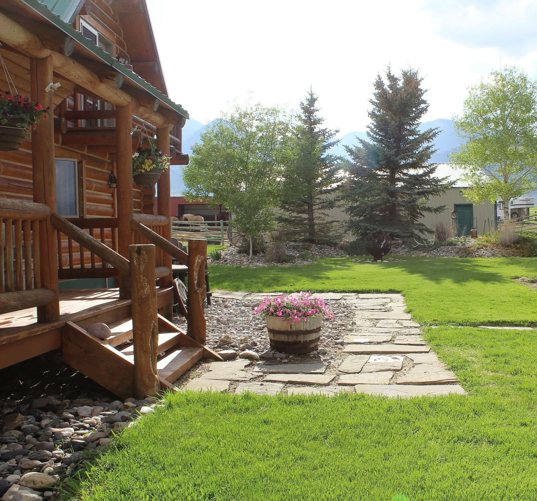 A rustic log cabin with a wooden porch overlooks a green lawn, stone pathway, and trees. Perfect as a ranch for sale, it features a wooden barrel planter with pink flowers near the steps while sunlight brightens the scene.