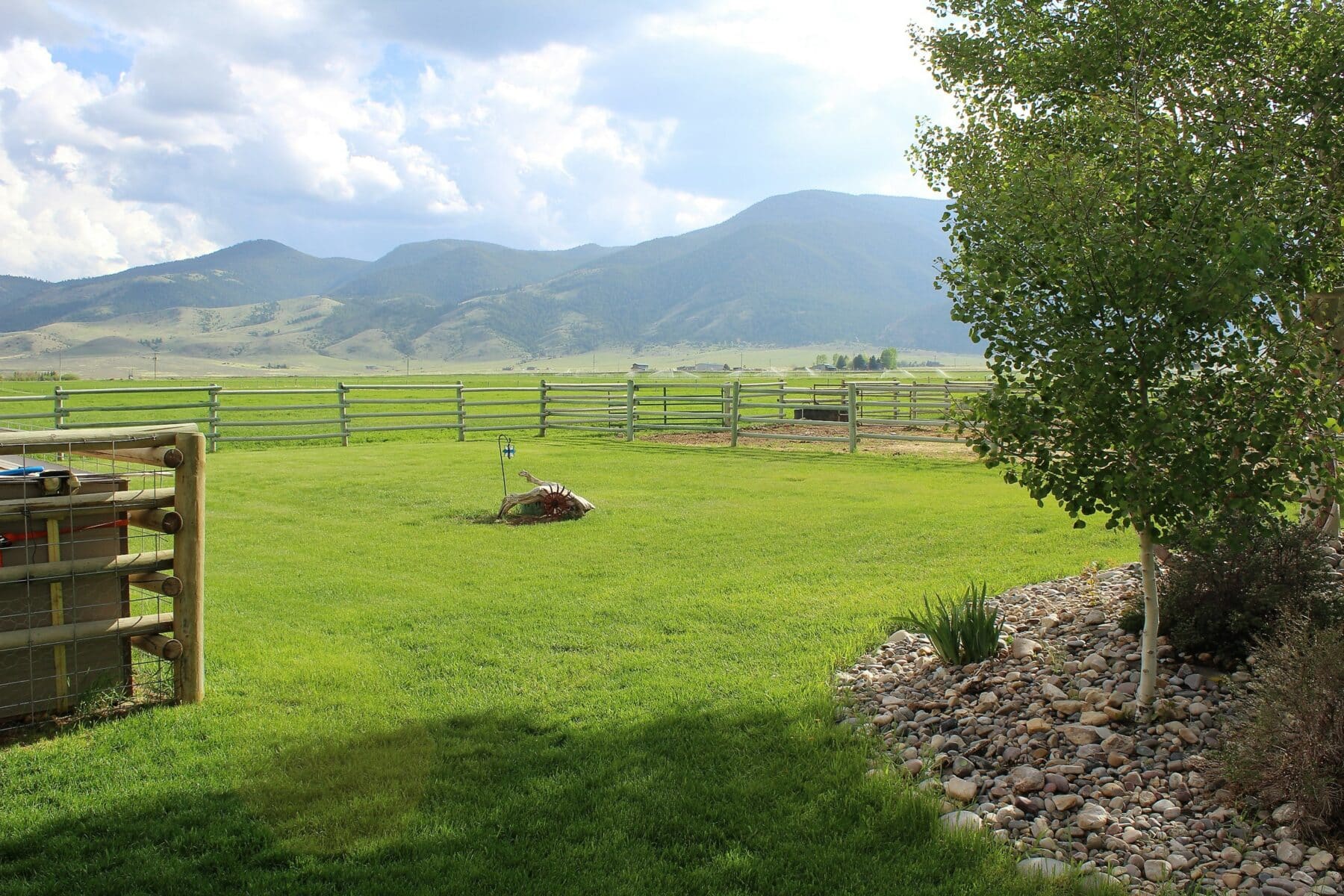 A green lawn with a tree and decorative rocks in the foreground, a small windmill and fence in the midground, and distant mountains under a partly cloudy sky—perfect recreational land or potential ranch for sale.