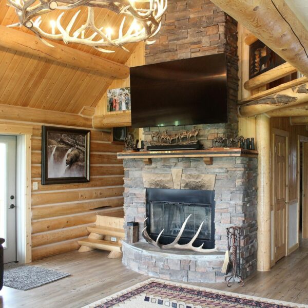 A cozy log cabin living room featuring a stone fireplace with antlers, a mounted TV above, wooden beams, animal-themed décor, and warm natural wood walls—perfect for relaxing after exploring nearby hunting property or land for sale.