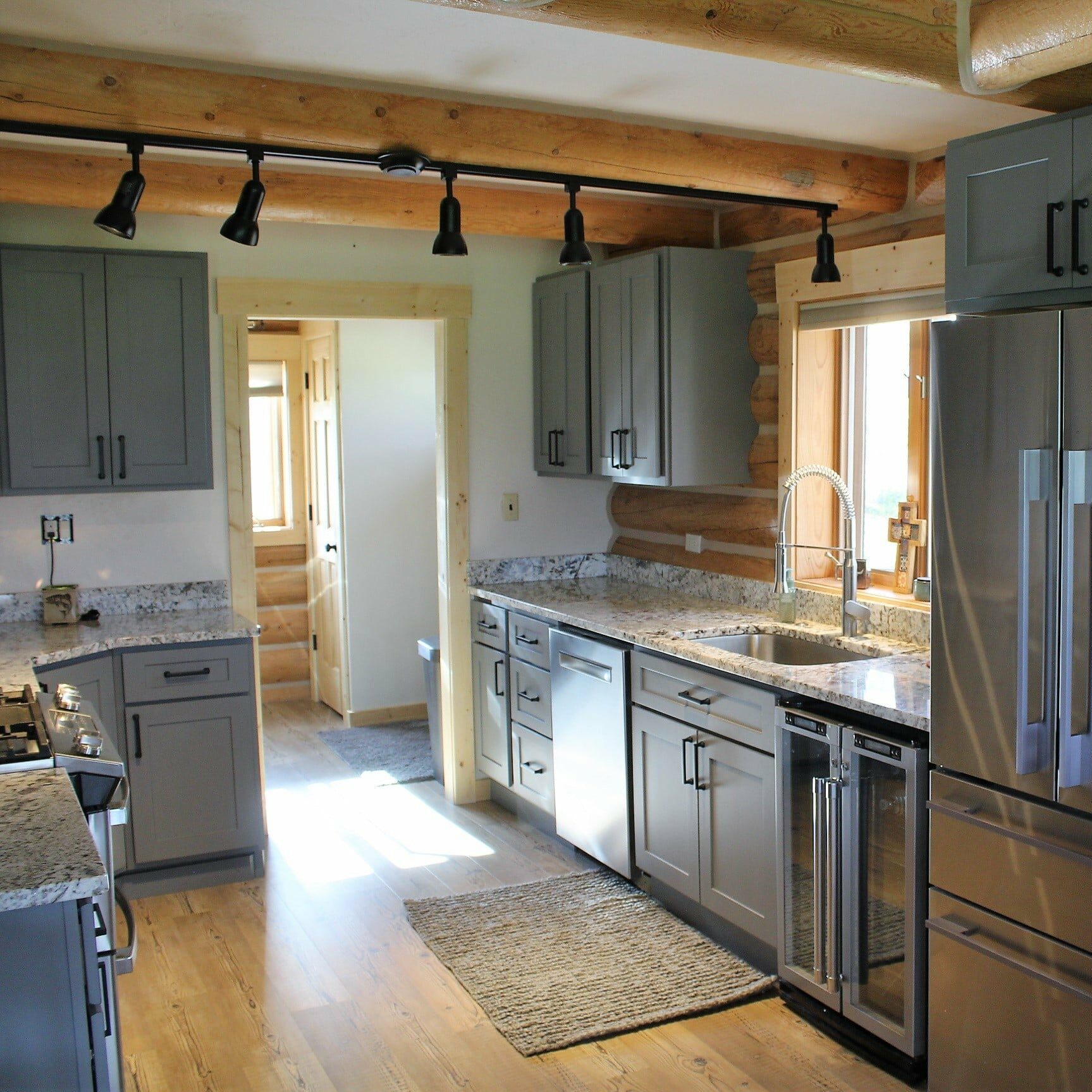 Modern kitchen with gray cabinets, stainless steel appliances, granite countertops, wood floors, exposed log beams, and large windows letting in natural light. Perfect for a cattle ranch or ranch for sale, with a rug near the sink area.