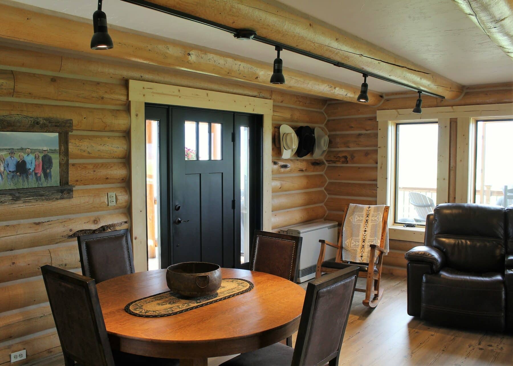 A cozy log cabin interior with wooden walls, a round dining table and four chairs, black front door, rocking chair, armchair, hats on the wall, large windows, and a framed picture—perfect for a cattle ranch or recreational land retreat.