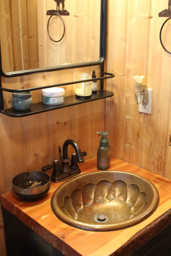 A rustic bathroom sink with a bronze basin, dark faucet, soap dispenser, and bowl on a wooden counter—perfect for a hunting property or recreational land. Two shelves above hold jars and a candle, set against wood-paneled walls and a mirror.