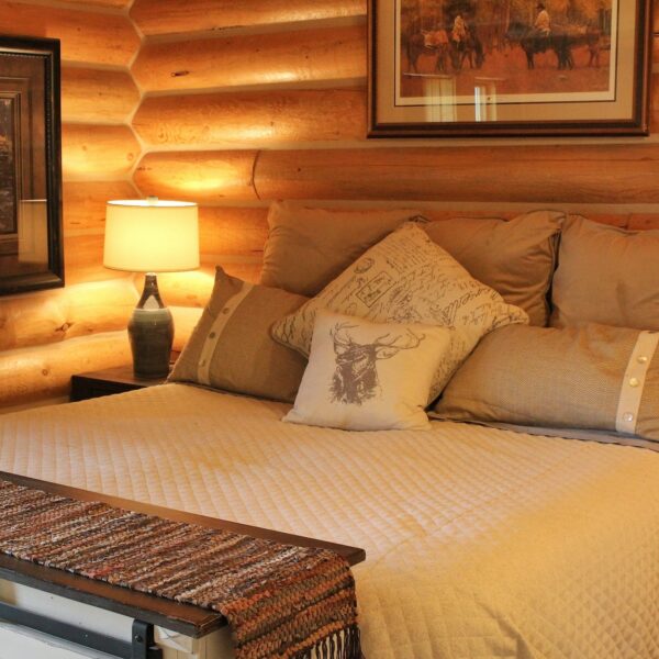 A cozy log cabin bedroom with wood walls, a neatly made bed with beige bedding and decorative pillows, a bedside table with a lamp, a woven bench at the foot of the bed, and framed art—perfect for relaxing after exploring your recreational land.
