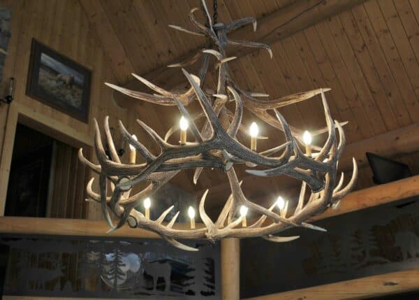A rustic chandelier made from antlers with glowing bulbs hangs from a wooden ceiling in a log cabin, surrounded by natural wood beams and outdoor-themed decor—perfect for a hunting property or cattle ranch.