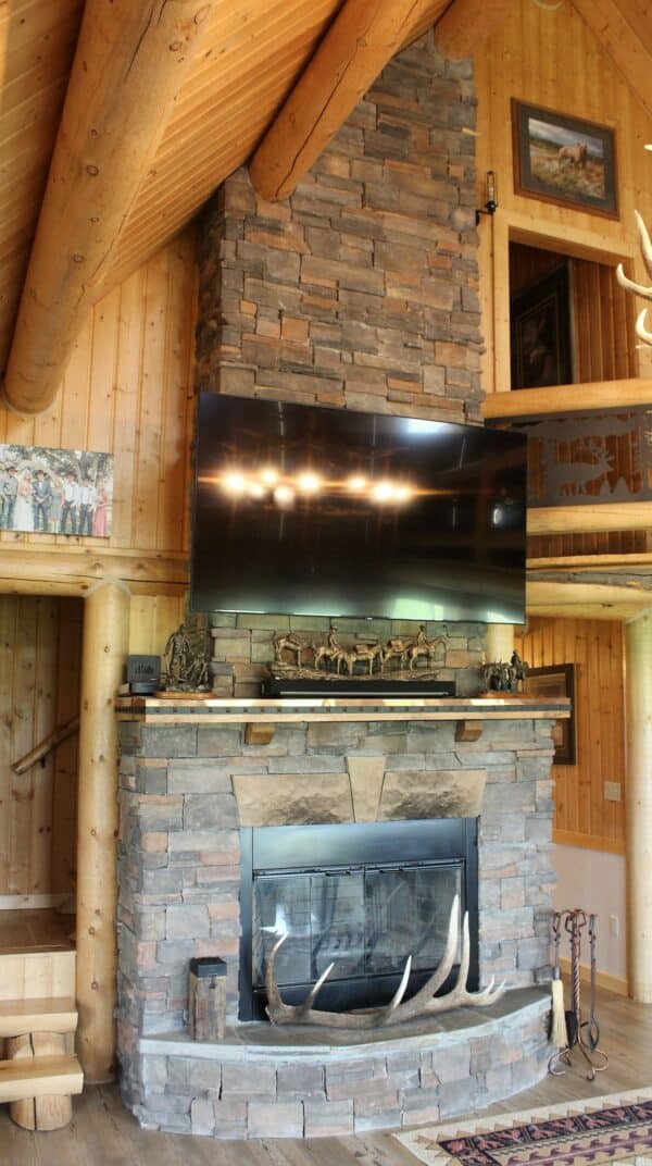 A rustic living room with a stone fireplace, antlers on the hearth, sculptures on the mantel, and a large TV mounted above. Wooden walls and log railings enhance the cozy cabin vibe—perfect for a hunting property or recreational land retreat.