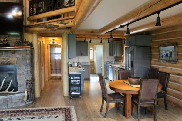 A cozy log cabin interior with wooden beams, a stone fireplace, dining table for four, modern kitchen appliances, and rustic decor throughout. Natural light fills the space, perfect for a ranch for sale or hunting property seekers.