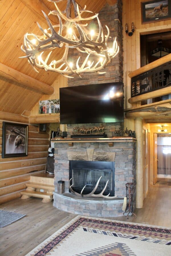 A rustic living room in a cozy cabin interior features log walls, a stone fireplace, antler chandelier, and a large TV above the mantel—perfect for a hunting property or recreational land. Animal-themed decor and wooden furniture complete the inviting space.