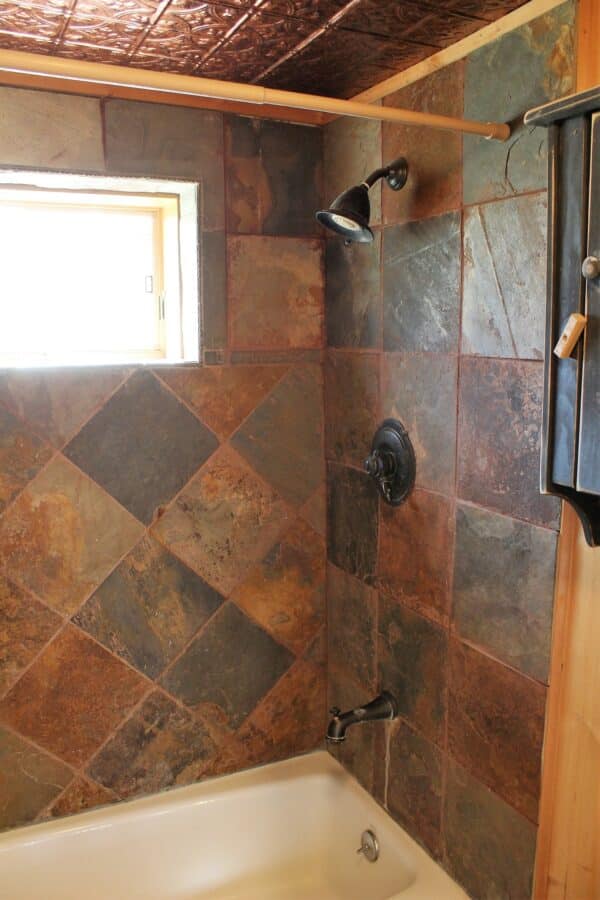 A bathtub and shower with dark, slate tile walls arranged in a diagonal pattern, featuring a black showerhead and faucet—perfect for relaxing after a day exploring your hunting property or cattle ranch. A small window lets in natural light.