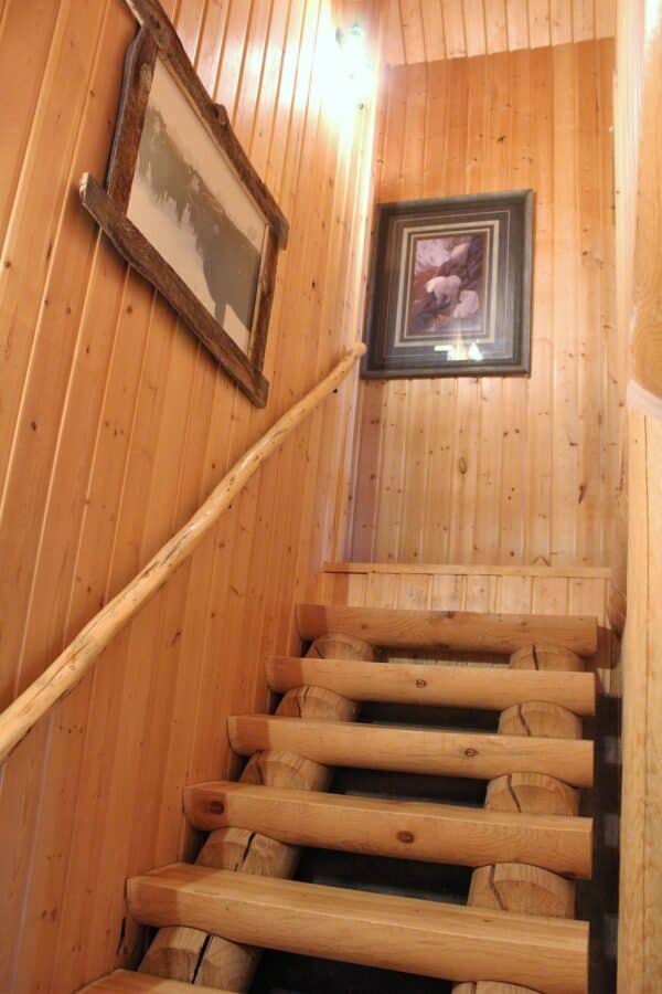 A rustic wooden staircase with log steps and a matching handrail leads up between pine-paneled walls, perfect for a cozy retreat on recreational land. Two framed pictures hang on the walls, and a light fixture illuminates the space from above.