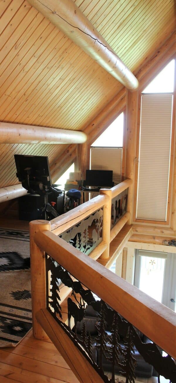 A cozy loft with wooden walls and ceiling, featuring two desks with monitors. The balcony railing is decorated with a cutout forest and wildlife design—ideal inspiration for those interested in hunting property or recreational land. Sunlight streams through tall windows.