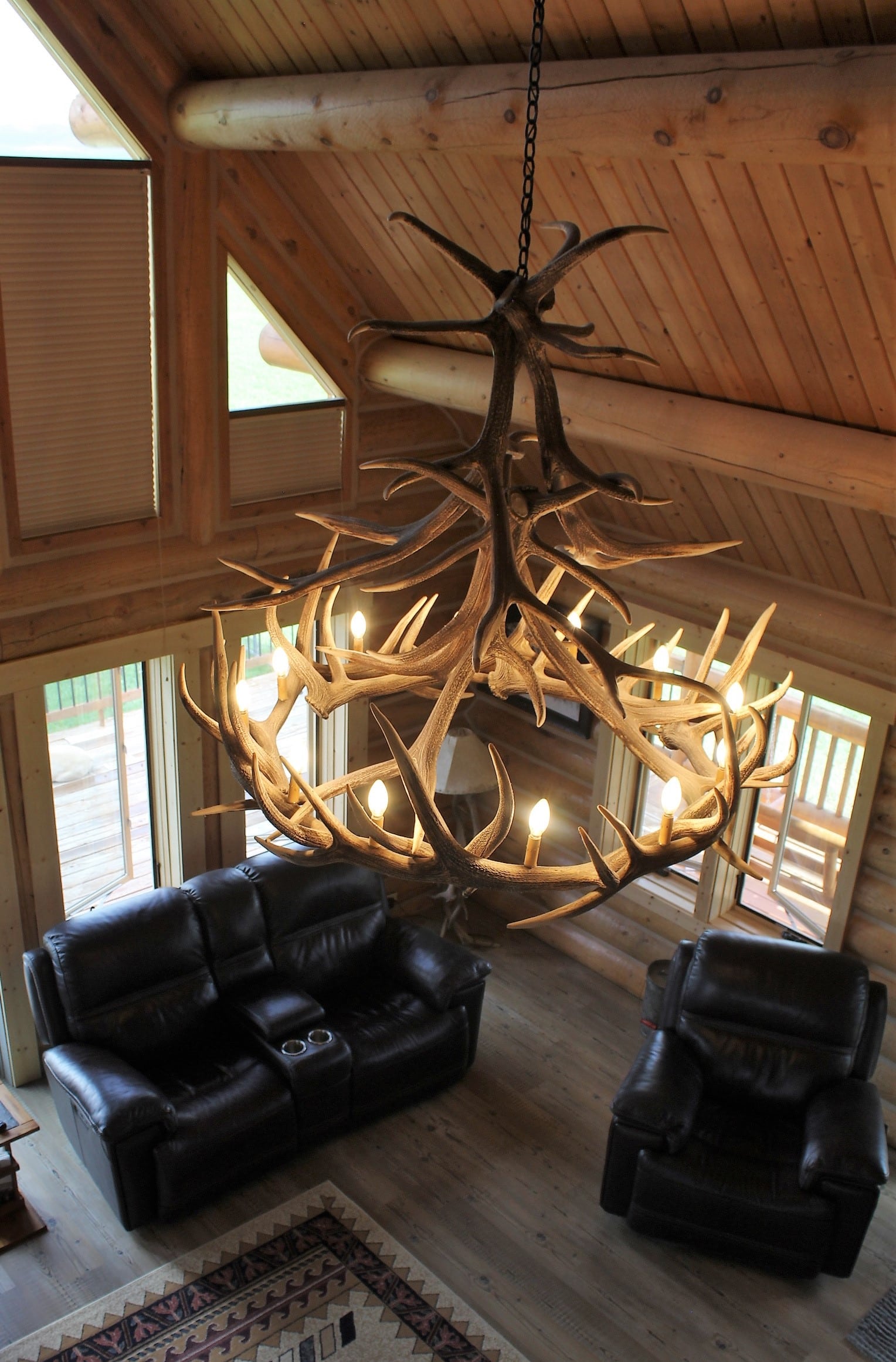 A large antler chandelier hangs from the wooden ceiling of a rustic living room, perfect for a cattle ranch, with leather sofas, wooden floors, and large windows letting in natural light.
