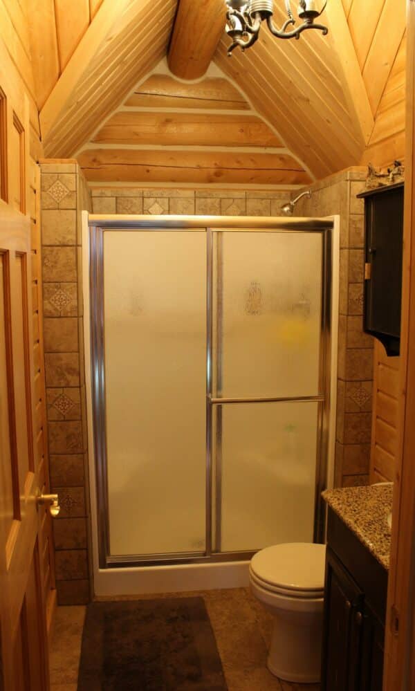 This cozy log cabin bathroom features wood-paneled walls and ceiling, a frosted glass shower, toilet, vanity with granite countertop, and a dark bath mat—perfect for a ranch for sale or recreational land retreat.