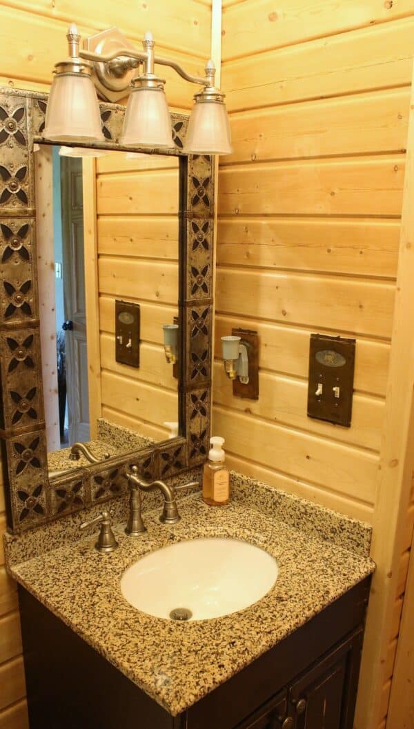 A rustic bathroom perfect for a hunting property, with wood-paneled walls, a granite countertop and built-in white sink, silver faucet, soap dispenser, decorative framed mirror, and a light fixture with three glass shades above the mirror.