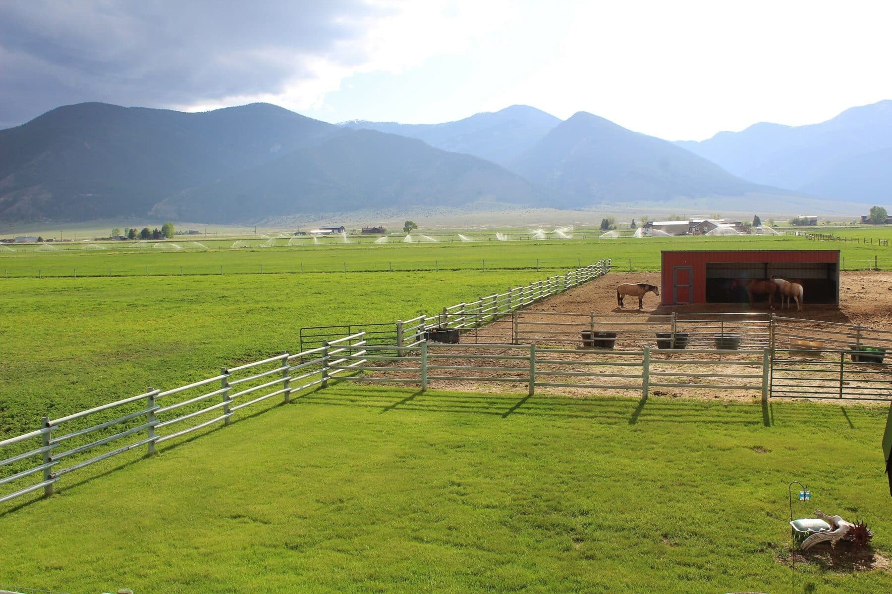 A fenced pasture with green grass and a dirt corral holds three horses and a red barn, set against mountains and a cloudy sky. Sprinklers water the field, making this scenic land for sale ideal as a hunting property or cattle ranch.