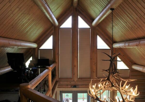 A cozy loft interior with wood-paneled walls and ceiling, triangular windows with blinds, a desk setup in the corner, and a rustic antler chandelier—perfect for relaxing after exploring nearby recreational land.