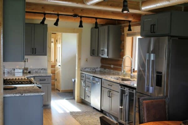 Modern kitchen with gray cabinets, stainless steel appliances, marble countertops, a large refrigerator, gas stove, and wooden floors. Natural light streams through a window above the sink; wooden beams on the ceiling give this ranch for sale rustic charm.