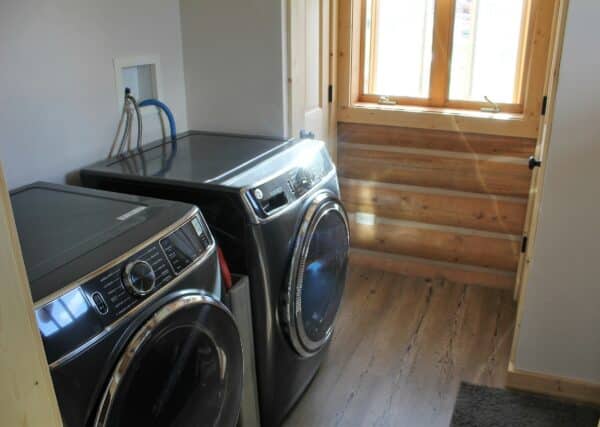 A small laundry room with a modern black washer and dryer, wood trim around a window, natural light, and wood plank flooring—ideal for a ranch for sale. Laundry hookups and a gray rug are also visible.