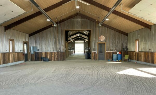 A spacious, clean barn interior with high wooden ceilings at a cattle ranch. Large open doors at each end, a clock on the wall, and various utility items along the right wall. Natural light streams in.