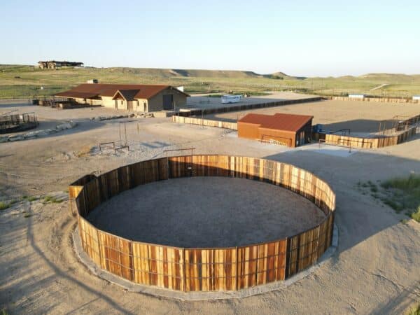 Aerial view of a cattle ranch with wooden buildings, a large circular corral, and fenced areas, set in a dry, open landscape with rolling hills in the background—ideal land for sale.
