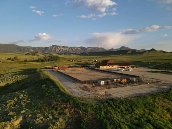 Aerial view of a large cattle ranch with fenced enclosures and buildings, set in a green, open landscape with mountains and a partly cloudy blue sky—ideal land for sale or use as a hunting property.