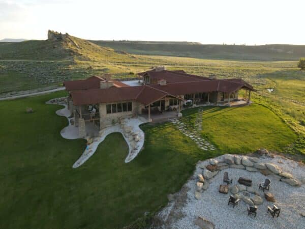 A sprawling ranch-style house with a red roof sits on green grass, surrounded by open fields. Ideal as a hunting property, it features a circular outdoor fire pit area with chairs and stones, and a rocky hill rises in the background.