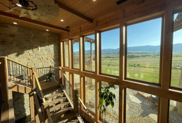 A sunlit stairway with wooden accents and large windows offers expansive views of green fields and distant mountains—ideal scenery for a ranch for sale. Indoor plants and stone walls add a rustic touch under the clear blue sky.