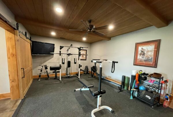 Home gym room with weightlifting equipment, bench, resistance bands, foam rollers, dumbbells, a TV on the wall, and a wooden sliding door—perfect for workouts before heading out onto your hunting property or cattle ranch. Framed picture and ceiling fan above.